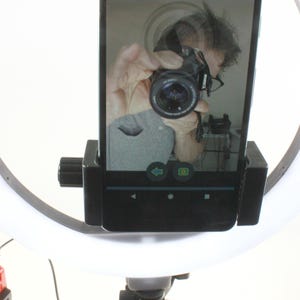 May include: A white ring light with a black phone holder attached to it. The phone is displaying a selfie of a person holding a camera. The ring light is mounted on a black stand with a silver base.