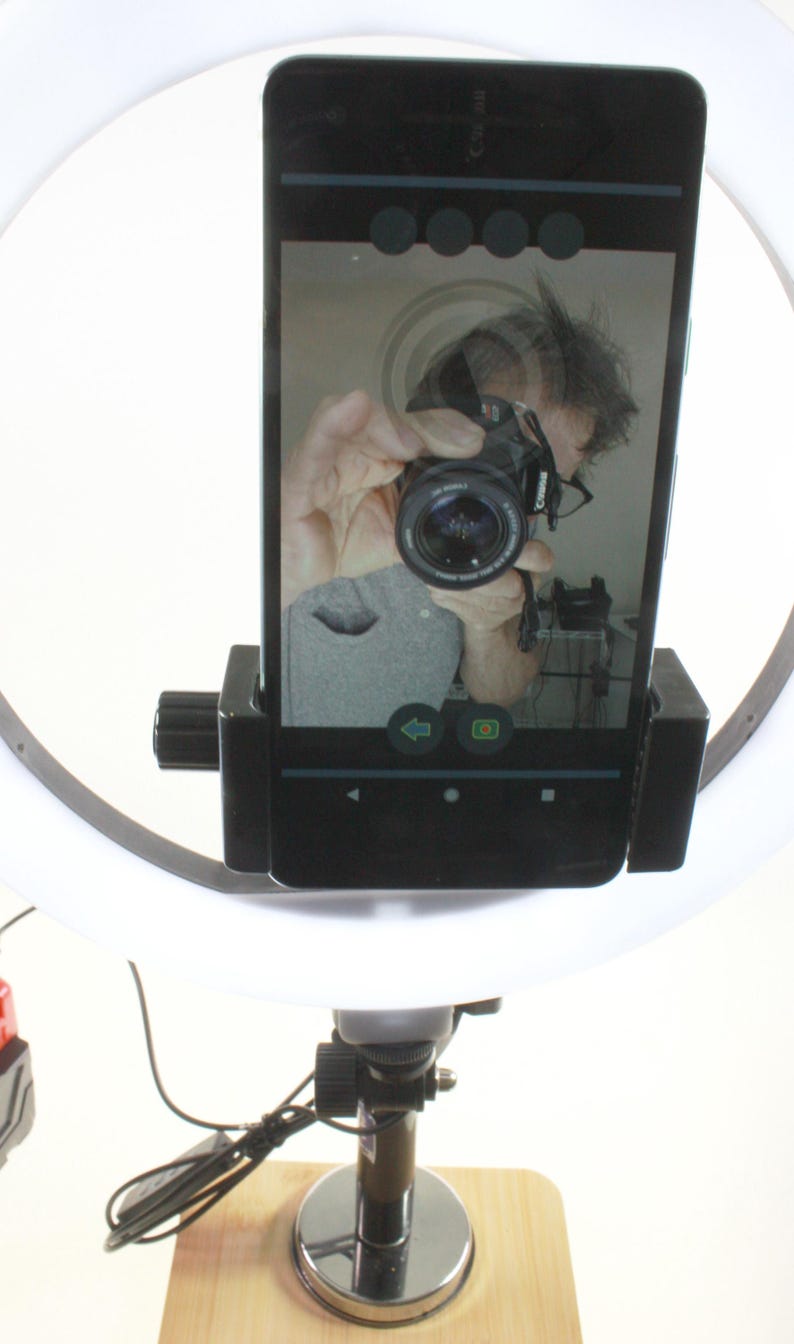 May include: A white ring light with a black phone holder attached to it. The phone is displaying a selfie of a person holding a camera. The ring light is mounted on a black stand with a silver base.