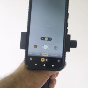 May include: A black smartphone with a protective case is held in a black grip. The phone's screen displays a camera interface with options for photo, portrait, and night sight modes. The phone is mounted in a holder.