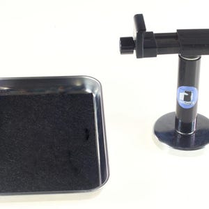 May include: A black and silver metal tray with a black foam insert sits next to a black stand with a circular base. The stand has a black adjustable arm and a circular base. The stand has a blue and white logo.