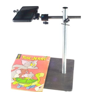 May include: A black smartphone is mounted on a silver metal stand, positioned above a vintage "Tom & Jerry" comic book. The comic book features a colorful illustration of the cartoon characters.
