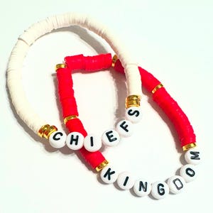 Könnte beinhalten: Zwei Armbänder mit weißen und roten Perlen. Das rote Armband hat goldene Perlen und weiße Buchstaben, die "CHIEFS" buchstabieren. Das weiße Armband hat goldene Perlen und weiße Buchstaben, die "KINGDOM" buchstabieren.