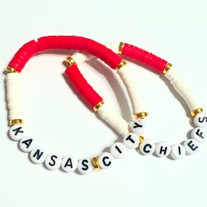 Könnte beinhalten: Zwei Armbänder mit weißen und roten Perlen und goldenen Akzenten. Die Perlen buchstabieren "Kansas City Chiefs".