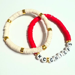 Könnte beinhalten: Zwei Armbänder, eines weiß und eines rot, mit goldenen Perlen und weißen Buchstabenperlen, die "K.C. CHIEFS" buchstabieren.