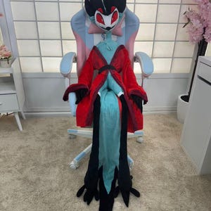 May include: A plush character costume featuring a red and black robe, a teal torso, and a large head with a white face, black accents, and red eyes. The costume is seated in a pink and blue gaming chair.