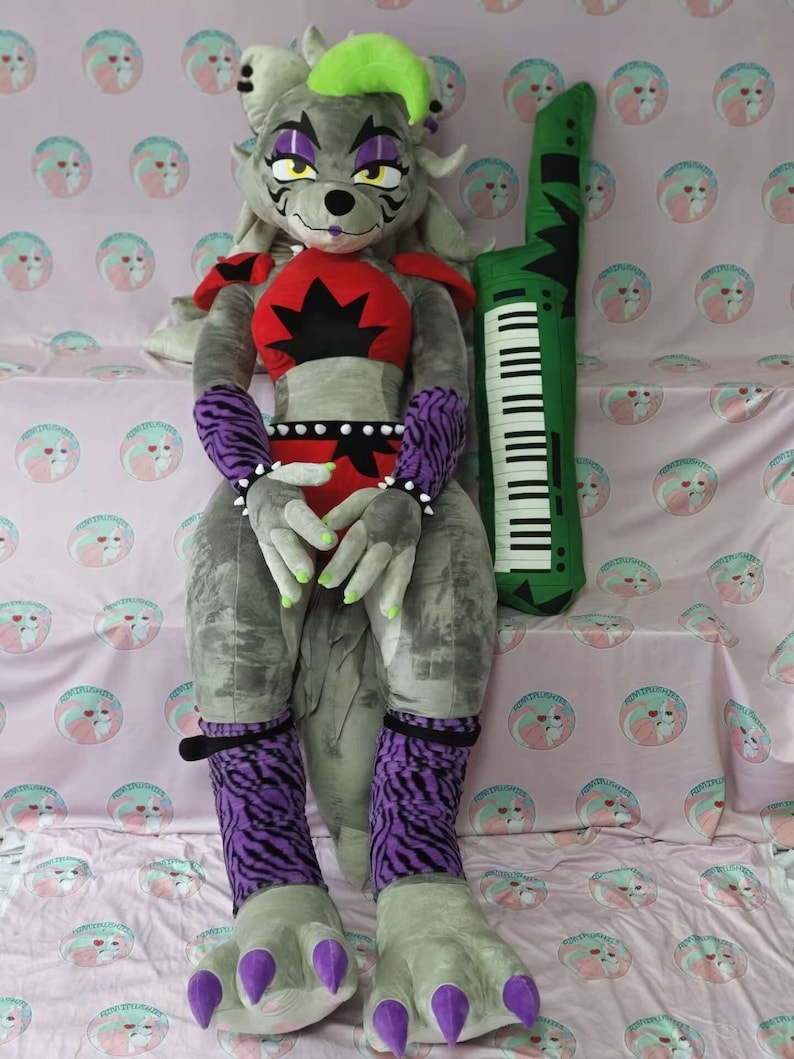 May include: A large gray plush toy with purple and black striped sleeves and leggings. The toy has a green mohawk and is holding its hands together. The toy is sitting on a pink and white patterned fabric with a green and black keyboard plush toy beside it.