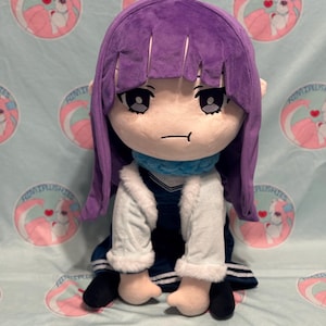 Può includere: Una bambola di peluche con una testa grande, capelli viola e una sciarpa blu. La bambola indossa una giacca bianca e una gonna blu scuro. La bambola ha un'espressione neutra ed è seduta. Lo sfondo è un tessuto azzurro con loghi rosa e bianchi ripetuti.