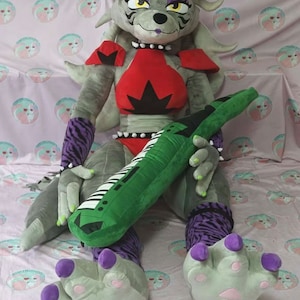 May include: A large gray plush wolf with a green mohawk, wearing a red and black outfit, holding a green keyboard-shaped pillow. The wolf has purple claws and is sitting on a pink and white patterned fabric.