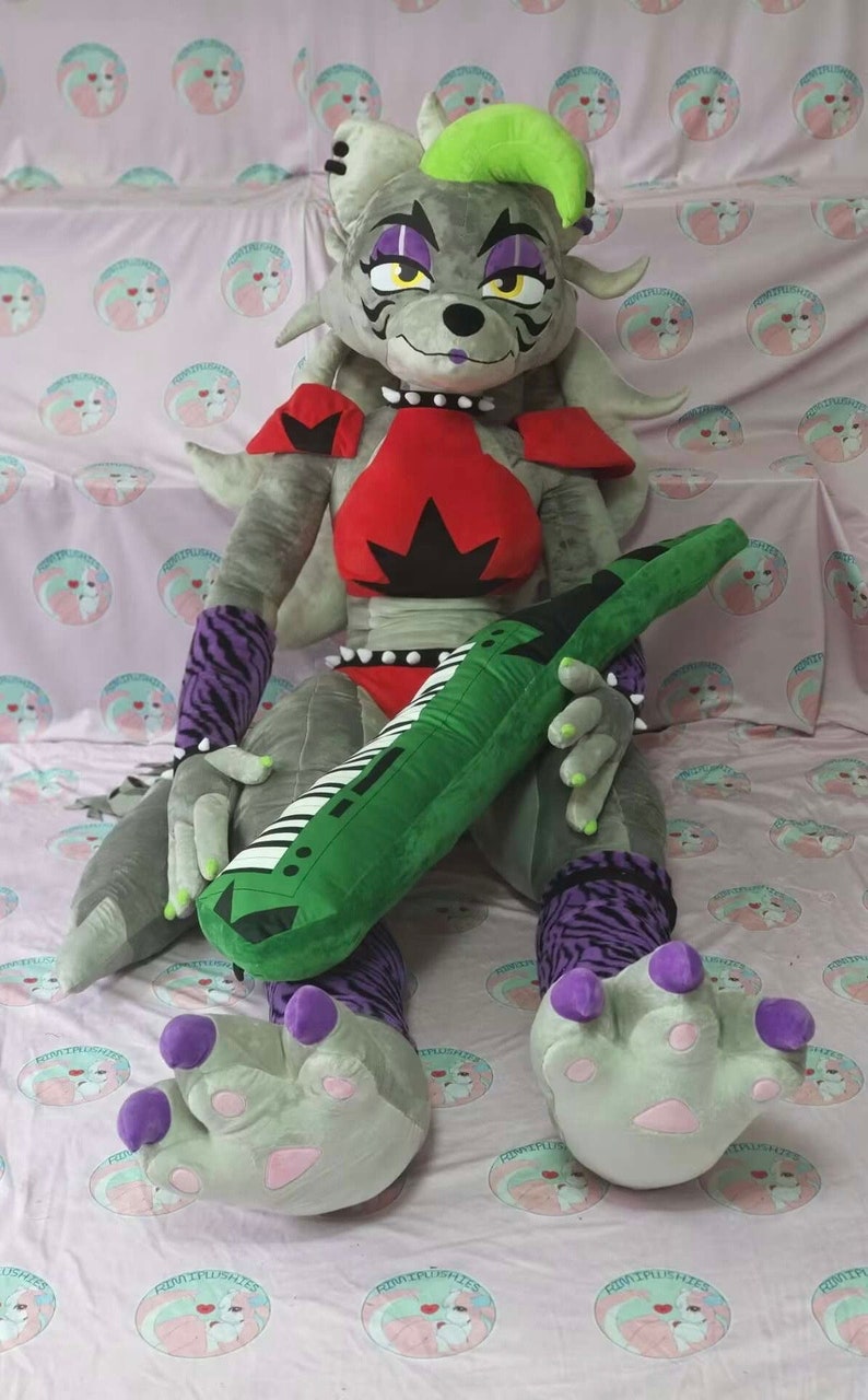 May include: A large gray plush wolf with a green mohawk, wearing a red and black outfit, holding a green keyboard-shaped pillow. The wolf has purple claws and is sitting on a pink and white patterned fabric.