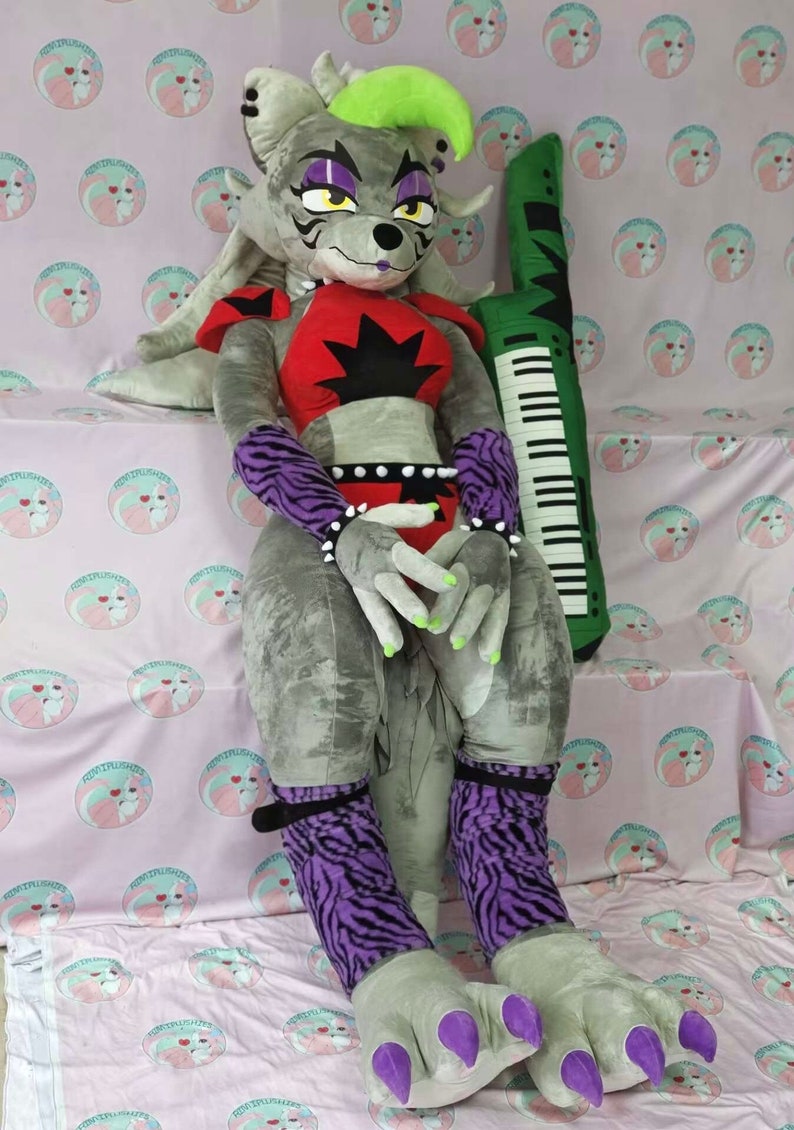 May include: A large gray plush toy with purple and black striped socks, green claws, and a red and black top. The toy is sitting on a pink and white patterned fabric with a green keyboard pillow behind it.