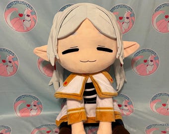 May include: A plush doll with a large head, long ears, and closed eyes. The doll wears a white and gold outfit with brown boots. The background is a light blue fabric with a repeating pattern of a pink and white cartoon character and the text "RIMIPLUSHIES".
