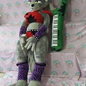 May include: A large gray plush toy with purple and black striped leg warmers, green hair, and red and black clothing. The toy is sitting on a pink and white patterned blanket with a green keyboard pillow beside it.