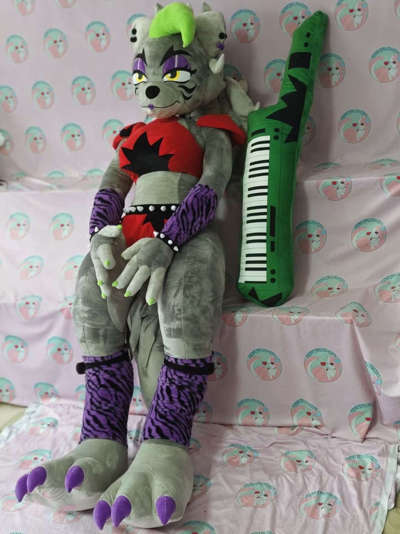 May include: A large gray plush toy with purple and black striped leg warmers, green hair, and red and black clothing. The toy is sitting on a pink and white patterned blanket with a green keyboard pillow beside it.