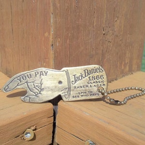 May include: A vintage-style bottle opener with a brass finish. The opener is shaped like a hand pointing, with the words "YOU PAY" and "Jack Daniel's 1866 Classic Amber Lager" printed on it. It includes a small chain.