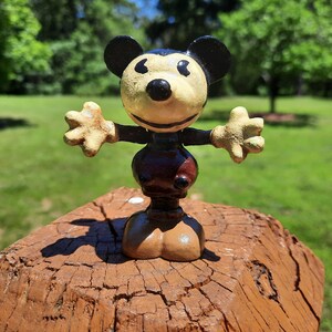 May include: A vintage Mickey Mouse figurine with outstretched arms. The figure has a cream-coloured face, black ears, and a black nose. The body is dark brown with a lighter brown base. The figurine is displayed on a weathered, brown wooden surface.