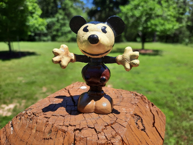 May include: A vintage Mickey Mouse figurine with outstretched arms. The figure has a cream-coloured face, black ears, and a black nose. The body is dark brown with a lighter brown base. The figurine is displayed on a weathered, brown wooden surface.