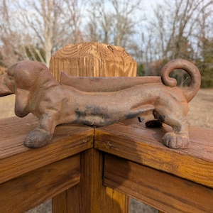 May include: A brown cast iron dachshund dog figurine. The dog is lying down with its head up and tail curled. The figurine is on a wooden deck railing. The dog is approximately 12 inches long.