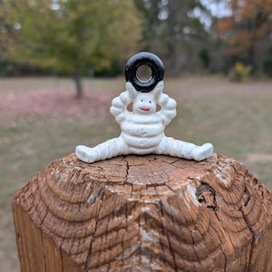 Könnte beinhalten: Eine weiße, segmentierte, reifenartige Figur mit einem schwarzen Ring auf dem Kopf, roten Lippen sowie ausgestreckten Armen und Beinen. Die Figur sitzt mit weit gespreizten Beinen und weist eine texturierte, matte Oberfläche auf.