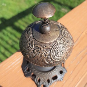 K&ouml;nnte beinhalten: Eine antike Messingglocke mit einem verzierten Blumendesign. Die Glocke hat einen federbelasteten Kl&ouml;ppel und steht auf einem dekorativen Sockel. Die Glocke ist ein Vintage-Artikel, wahrscheinlich aus dem fr&uuml;hen 20. Jahrhundert.