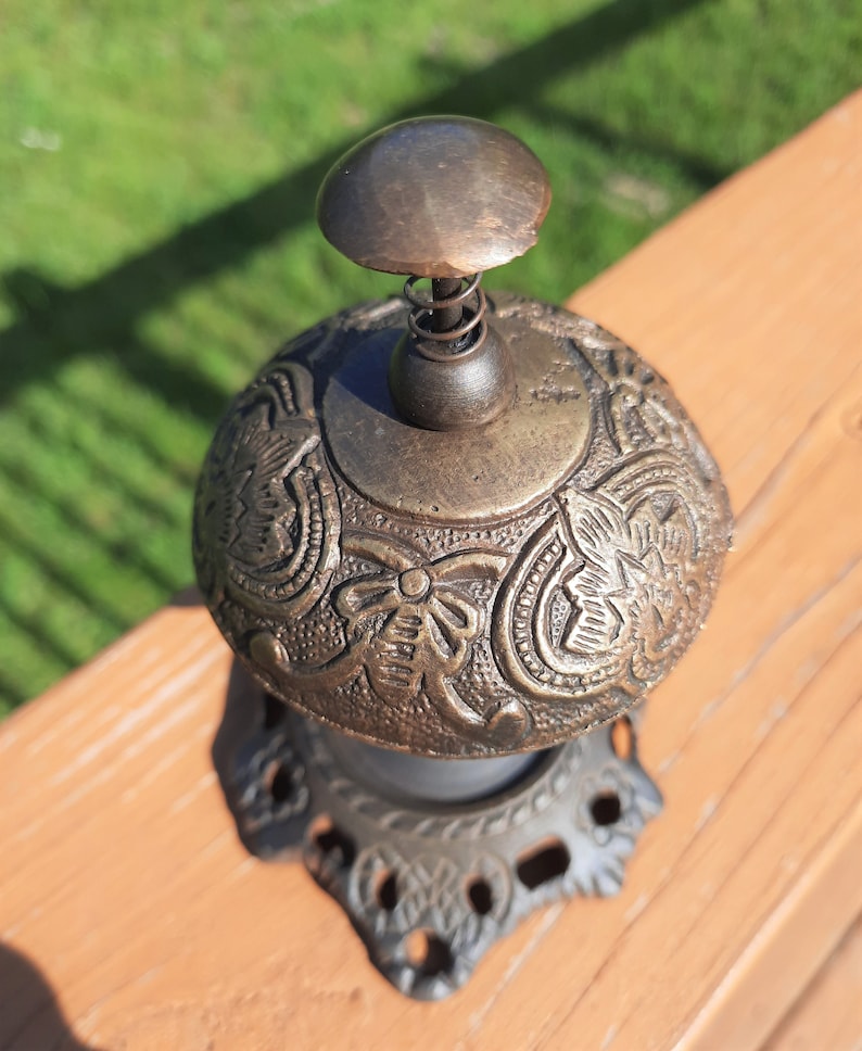 K&ouml;nnte beinhalten: Eine antike Messingglocke mit einem verzierten Blumendesign. Die Glocke hat einen federbelasteten Kl&ouml;ppel und steht auf einem dekorativen Sockel. Die Glocke ist ein Vintage-Artikel, wahrscheinlich aus dem fr&uuml;hen 20. Jahrhundert.