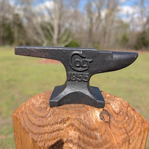 May include: A small, black anvil with the word "COLT" and the year "1855" stamped on its side. The anvil is set on a wooden post, with a blurred green and brown background.