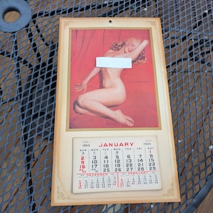 Könnte beinhalten: Ein alter Wandkalender von 1955 mit einer Pin-up-Illustration einer blonden Frau vor rotem Hintergrund. Der Kalender zeigt den Monat Januar mit rotem und schwarzem Text sowie kleine Kalender für Dezember und Februar.