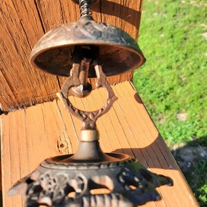 K&ouml;nnte beinhalten: Antike bronzefarbene Schreibtischglocke mit dekorativem Sockel und federbelastetem Kl&ouml;ppel. Die Glocke hat eine verzierte, gew&ouml;lbte Oberseite und einen detaillierten Griff. Der Sockel weist ein florales Design auf. Die Glocke steht auf einer Holzoberfl&auml;che.