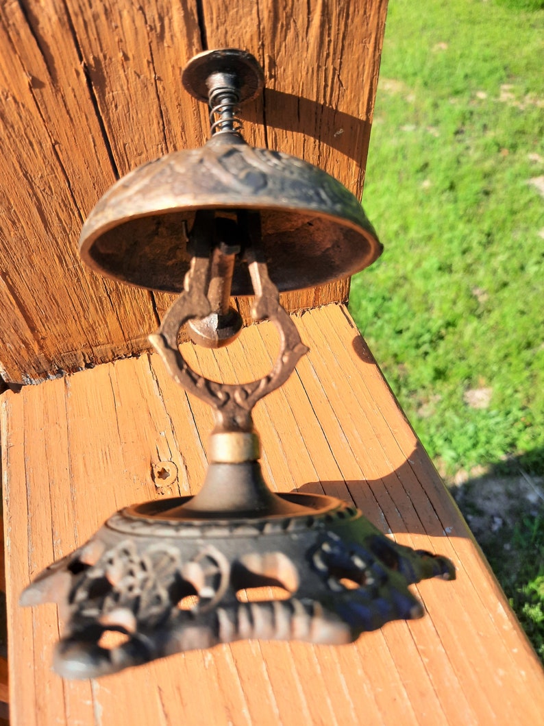 K&ouml;nnte beinhalten: Antike bronzefarbene Schreibtischglocke mit dekorativem Sockel und federbelastetem Kl&ouml;ppel. Die Glocke hat eine verzierte, gew&ouml;lbte Oberseite und einen detaillierten Griff. Der Sockel weist ein florales Design auf. Die Glocke steht auf einer Holzoberfl&auml;che.