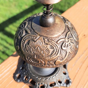 K&ouml;nnte beinhalten: Zierliche Glocke in Bronzeoptik mit floralen und sternf&ouml;rmigen Mustern. Sie hat eine Feder und einen kleinen Griff. Der Sockel ist dekorativ und durchbrochen. Die Glocke steht auf einer Holzoberfl&auml;che.