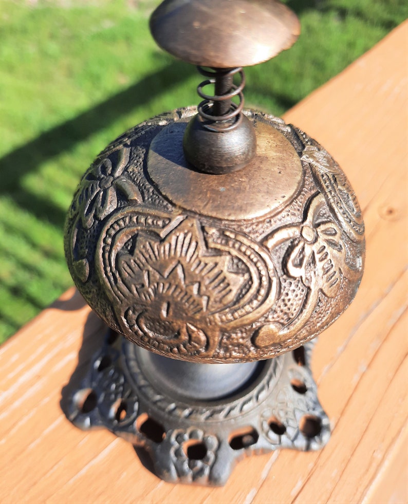 K&ouml;nnte beinhalten: Zierliche Glocke in Bronzeoptik mit floralen und sternf&ouml;rmigen Mustern. Sie hat eine Feder und einen kleinen Griff. Der Sockel ist dekorativ und durchbrochen. Die Glocke steht auf einer Holzoberfl&auml;che.