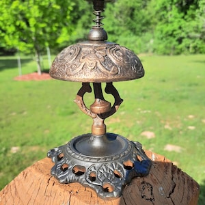 K&ouml;nnte beinhalten: Eine Schreibtischglocke im antiken Stil mit Bronze-Finish. Die Glocke hat eine verzierte, kuppelf&ouml;rmige Oberseite mit gepr&auml;gten floralen Mustern, einen Federmechanismus und einen dekorativen Sockel. Die Glocke ist etwa 15 cm hoch.