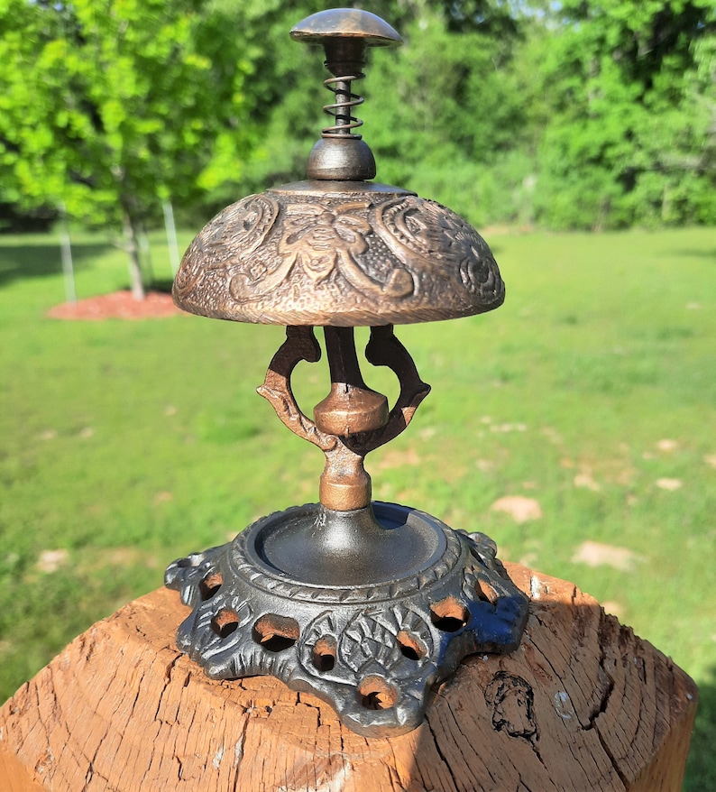 K&ouml;nnte beinhalten: Eine Schreibtischglocke im antiken Stil mit Bronze-Finish. Die Glocke hat eine verzierte, kuppelf&ouml;rmige Oberseite mit gepr&auml;gten floralen Mustern, einen Federmechanismus und einen dekorativen Sockel. Die Glocke ist etwa 15 cm hoch.