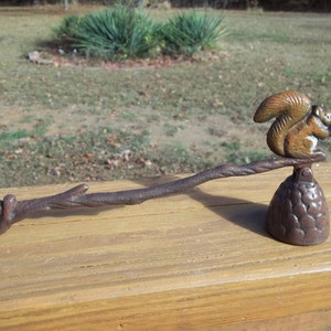 May include: A cast iron candle snuffer with a squirrel design. The snuffer has a long, twig-like handle and a bell-shaped base. The squirrel is sitting on the handle and holding a nut in its paws.