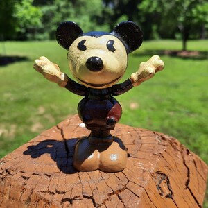 May include: A vintage Mickey Mouse figurine with arms outstretched. The figure has a black head and ears, a tan face and hands, and a dark brown body with a red accent. It stands on a weathered wooden surface against a blurred green background.