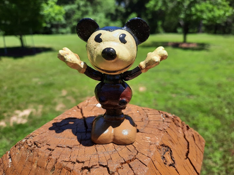 May include: A vintage Mickey Mouse figurine with arms outstretched. The figure has a black head and ears, a tan face and hands, and a dark brown body with a red accent. It stands on a weathered wooden surface against a blurred green background.