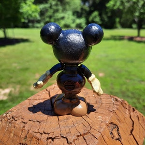 May include: A black Mickey Mouse figurine with a round body, large ears, and beige gloves and shoes. The figure is standing on a brown wooden surface against a backdrop of green grass and trees.