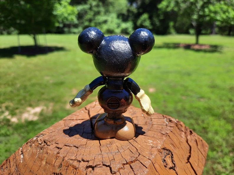 May include: A black Mickey Mouse figurine with a round body, large ears, and beige gloves and shoes. The figure is standing on a brown wooden surface against a backdrop of green grass and trees.