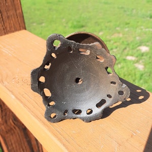 K&ouml;nnte beinhalten: Ein dunkles Metallobjekt im antiken Stil mit einem dekorativen, durchbrochenen Design. Das Objekt hat eine kreisf&ouml;rmige Basis und einen detaillierten, gezackten Rand mit zahlreichen kleinen L&ouml;chern. Das Metall hat ein verwittertes Aussehen.