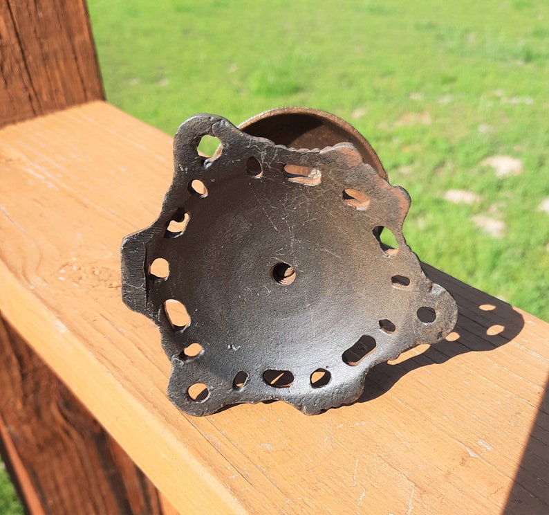 K&ouml;nnte beinhalten: Ein dunkles Metallobjekt im antiken Stil mit einem dekorativen, durchbrochenen Design. Das Objekt hat eine kreisf&ouml;rmige Basis und einen detaillierten, gezackten Rand mit zahlreichen kleinen L&ouml;chern. Das Metall hat ein verwittertes Aussehen.