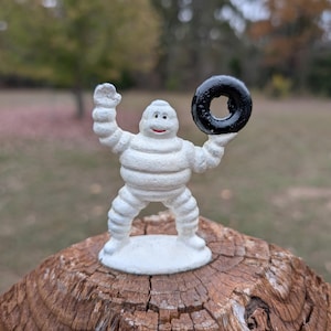 Könnte beinhalten: Eine weiße Michelin-Männchen-Figur auf einem runden Sockel. Die Figur hat einen strukturierten, reifenartigen Körper, ein lächelndes Gesicht mit roten Lippen und einen erhobenen Arm. Die andere Hand hält ein kleines, schwarzes, reifenförmiges Objekt.