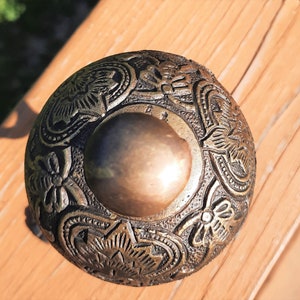 K&ouml;nnte beinhalten: Ein detailliertes Metallobjekt im antiken Stil mit einem gepr&auml;gten Blumendesign. Der zentrale Bereich ist eine glatte, kreisf&ouml;rmige Form, umgeben von komplizierten Mustern. Das Objekt hat eine bronzefarbene Farbe und scheint aus Metall zu sein.