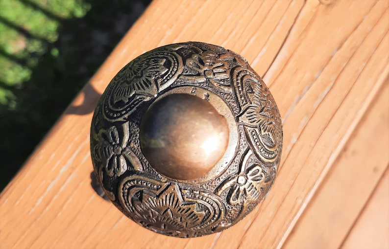 K&ouml;nnte beinhalten: Ein detailliertes Metallobjekt im antiken Stil mit einem gepr&auml;gten Blumendesign. Der zentrale Bereich ist eine glatte, kreisf&ouml;rmige Form, umgeben von komplizierten Mustern. Das Objekt hat eine bronzefarbene Farbe und scheint aus Metall zu sein.