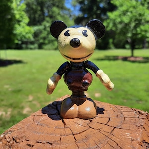May include: A vintage Mickey Mouse figurine with a cream face, black ears, and a brown body. The figure has articulated arms and legs, and is standing on a brown wooden surface. The background is a blurred green lawn and trees.