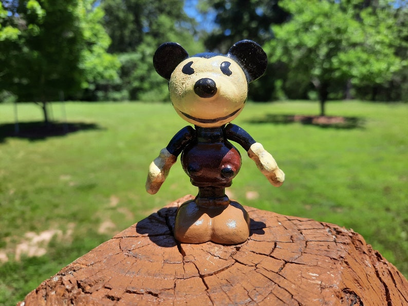May include: A vintage Mickey Mouse figurine with a cream face, black ears, and a brown body. The figure has articulated arms and legs, and is standing on a brown wooden surface. The background is a blurred green lawn and trees.