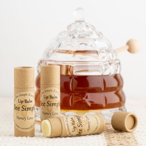 May include: Three brown cardboard tubes with labels that read "Bee Simple 2 on 1 Lip Balm Honey Love" and contain a white lip balm. The tubes are sitting on a beige surface in front of a glass honey pot with a wooden dipper.