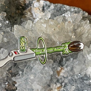 May include: A silver and green enamel pin depicting a cartoon device shooting a green beam of energy towards a brown target. The device has a red button and a small green container with a green object inside.