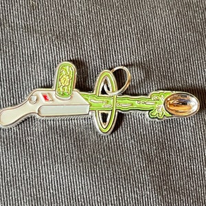 May include: A silver-colored metal pin depicting a cartoon-style device with a green goo-like substance shooting out of it. The device has a red button and a green container on top. The goo is shooting into a silver spoon.