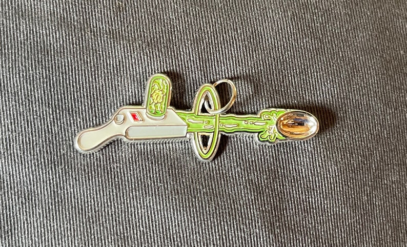 May include: A silver-colored metal pin depicting a cartoon-style device with a green goo-like substance shooting out of it. The device has a red button and a green container on top. The goo is shooting into a silver spoon.