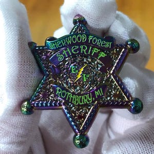 May include: A six-pointed star-shaped badge with iridescent colors of purple, green, and blue. The badge reads "Sherwood Forest Sheriff" and "Rothbury, MI" with a lightning bolt and the letters "EF" in the center.