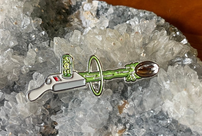 May include: A silver and green enamel pin depicting a cartoon device shooting a green beam of energy towards a brown target. The device has a red button and a small green container with a green object inside.